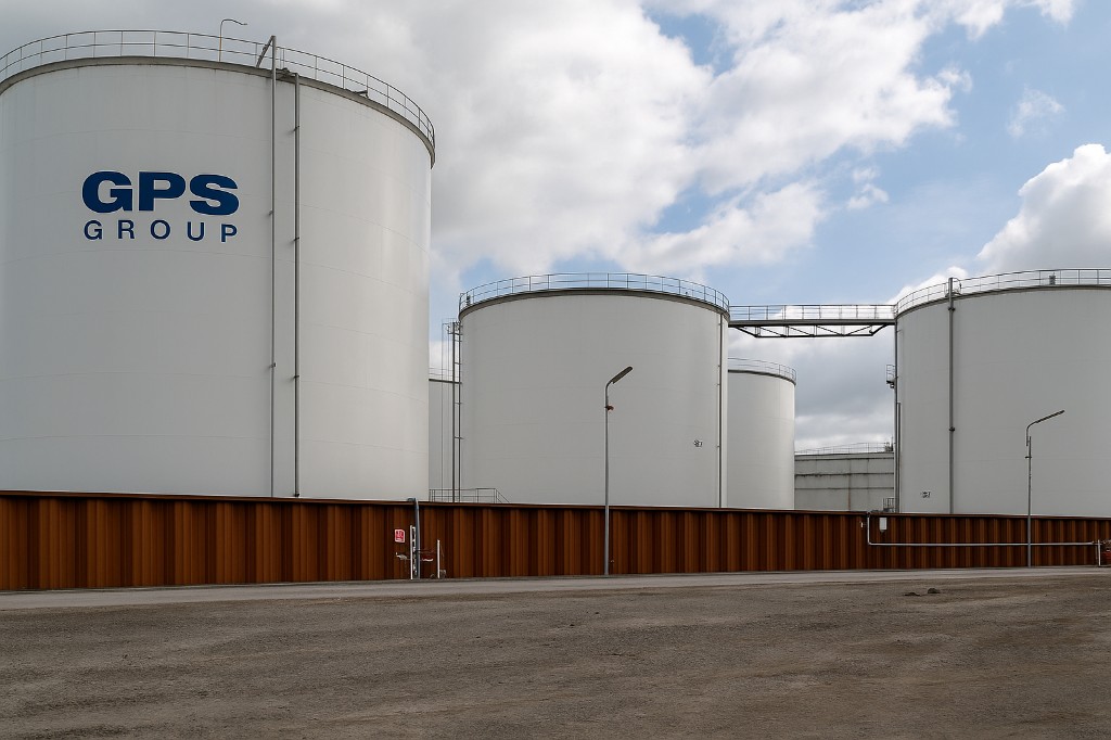 White storage tanks, the nearest marked GPS Group in blue, behind a rust-colored wall.